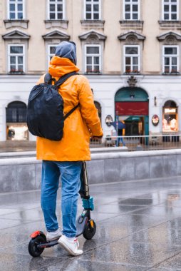 Sarı yağmurluklu adam elektrikli scooter fotokopi alanında şehir sokağının yanından geçiyor.