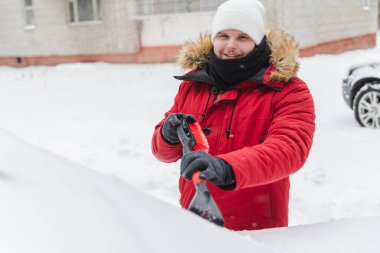 Kış mevsimi kar fırtınasından sonra kürk kapüşonlu bir adam araba temizliyor.