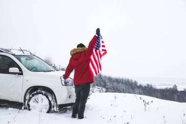 Tepenin tepesinde elinde ABD bayrağı olan bir adam arazi aracının yanında güzel bir kış manzarası arıyor. özgürlük kavramı