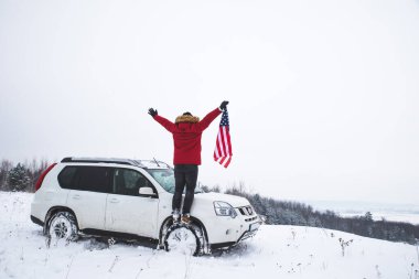 Tepenin tepesinde elinde ABD bayrağı olan bir adam arazi aracının yanında güzel bir kış manzarası arıyor. özgürlük kavramı