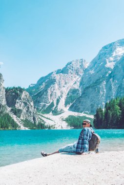 Arka planda sırt çantalı dağlarla gölün sahilinde oturan adam dolomitler.