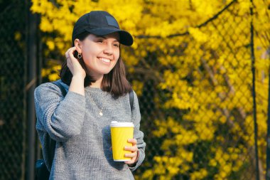 Sarı kağıt bardaktan açık havada kahve içen şapkalı genç ve şık bir kadın. Arkaplanda sarı çiçek açan çalılar