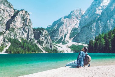 Arka planda sırt çantalı dağlarla gölün sahilinde oturan adam dolomitler.