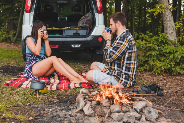 couple sitting on blanket near bonfire white suv vehicle on background hiking concept. woman taking picture on camera