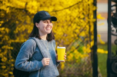Sarı kağıt bardaktan açık havada kahve içen şapkalı genç ve şık bir kadın. Arkaplanda sarı çiçek açan çalılar