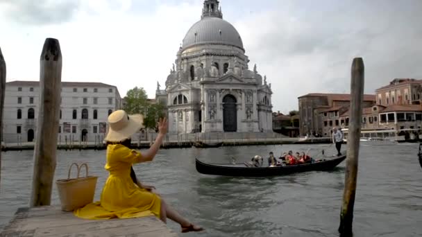 Женщина в желтом сарафане сидит на пирсе с видом на базилику Санта Мария делла Салют — стоковое видео