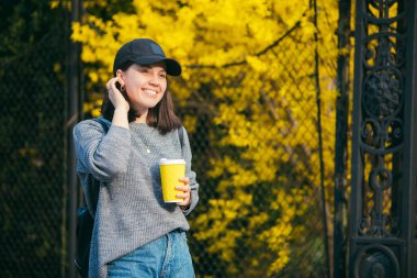 Sarı kağıt bardaktan açık havada kahve içen şapkalı genç ve şık bir kadın. Arkaplanda sarı çiçek açan çalılar