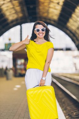Tren istasyonunda tekerlekli sarı bavullu, gülümseyen, çekici bir kadın.