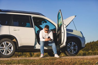 Gün batımında kaputu açık bir şekilde arabanın yanında oturan bir adam telefonla fotokopi uzayı hakkında konuşuyor.