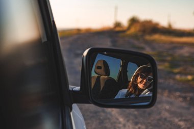 Güneş gözlüklü muhteşem kadın gün batımında araba aynasında yansıyor. Yol gezisi