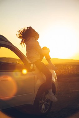 Kadın gün batımının tadını çıkarmak için yol kenarında duruyor. Araba kaputunda oturuyor. Boşluğu kopyala