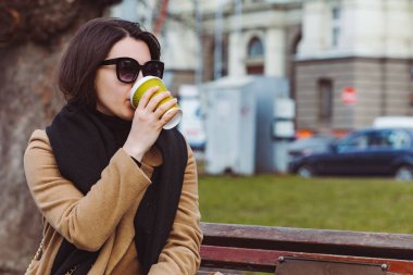 Güneş gözlüklü genç ve şık bir kadın bankta otururken kahve içer. yaşam tarzı konsepti