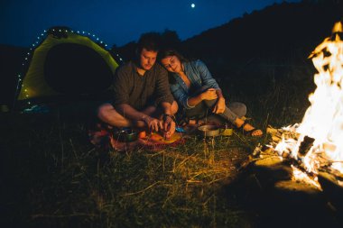 Gülümseyen neşeli çift şenlik ateşinin önünde yemek pişiriyor. kamp konsepti. Boşluğu kopyala