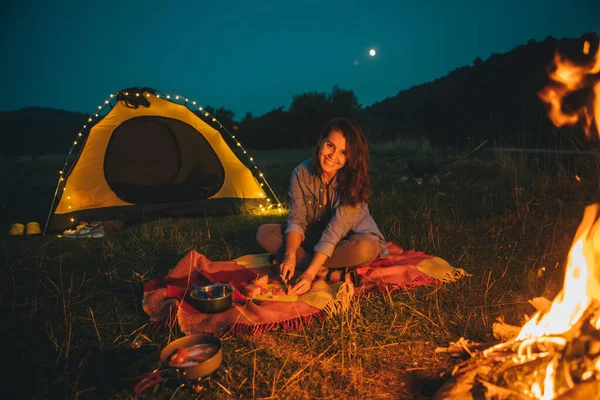 young pretty woman sitting on the ground in front of fire cooking dinner. yellow tent on background. copy space