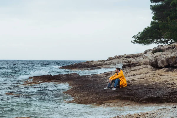 man sitting in yellow raincoat at rocky beach looking at stormy weather. copy space.