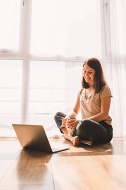 Yerde oturan kadın büyük pencerenin yanında dizüstü bilgisayarda çalışıyor. Kendini soyutlama. Karantinaya alındı. Telsiz çalışıyor. Görüntülü sohbet ile konuşmak