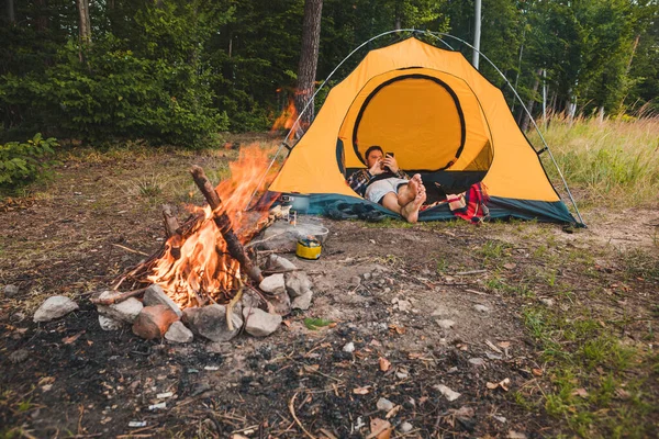man laying in tent with smartphone near campfire. hiking concept. travel concept