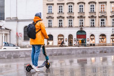 Sarı yağmurluklu adam elektrikli scooter fotokopi alanında şehir sokağının yanından geçiyor.