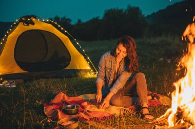 Genç ve güzel bir kadın ateşin önünde yemek pişiriyor. Arka planda sarı çadır. Boşluğu kopyala