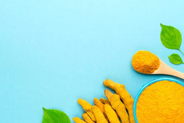 Mavi kasede ahşap kaşık, kurumuş kökler ve mavi arka planda yeşil yapraklarla zerdeçal tozu içeren düzen, boşluğu kopyala.