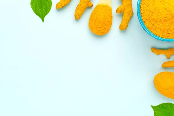 Mavi kasede ahşap kaşık, kurumuş kökler ve mavi arka planda yeşil yapraklarla zerdeçal tozu içeren düzen, boşluğu kopyala.