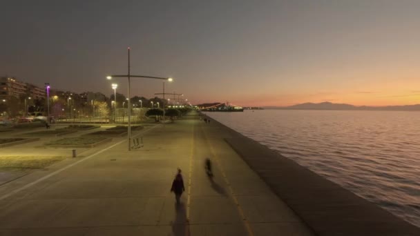 オレンジの日没の色を持つ都市のウォーターフロントで黄金の時間の経過を表示します ギリシャテッサロニキの改装された海岸沿いを歩く正体不明の群衆の夕日の時間表示 — ストック動画
