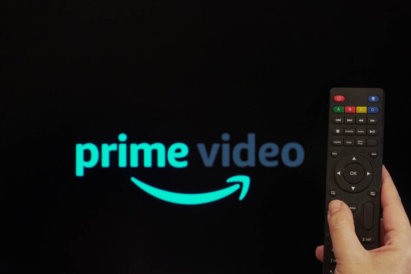 Thessaloniki, Greece - March 21 2020: Amazon streaming Service VoD content provider concept. Hand holds a multimedia remote control before a screen with Amazon Prime Video logo.