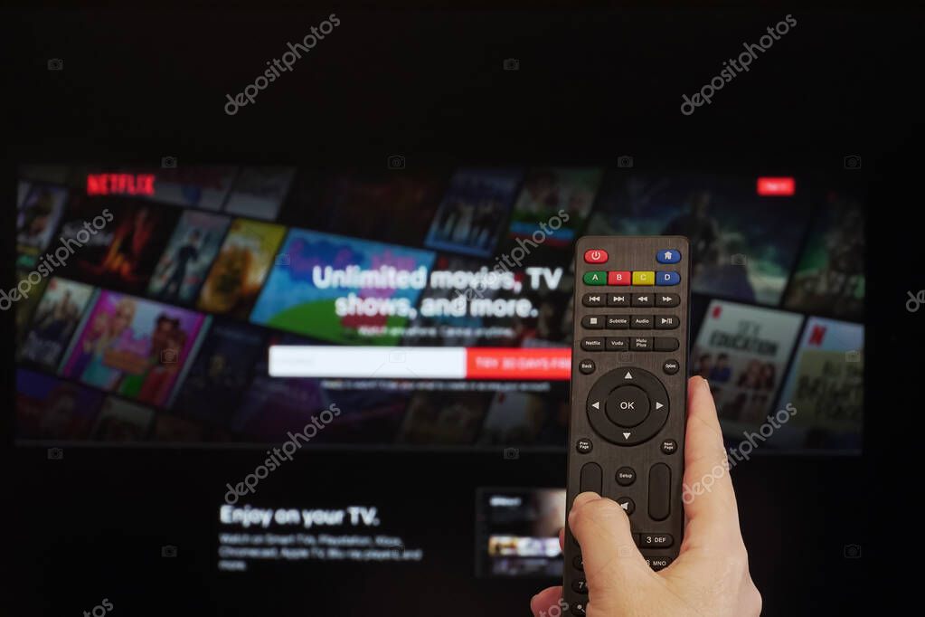 Thessaloniki, Greece - March 21 2020: Netflix streaming Service VoD content provider concept. Hand holds a multimedia remote control before a screen with Netflix app online watch login page.