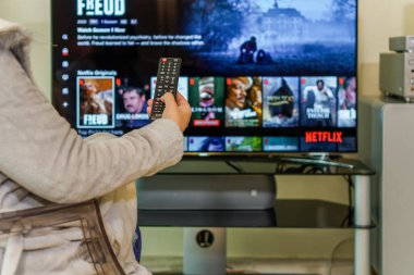 Selanik, Yunanistan - 4 Nisan 2020: TV Netflix canlı yayın hizmeti VoD içeriği. Elinde çokluortam uzaktan kumandası, geniş ekran yayın sağlayıcısı sayfasında görüntüsü olan günlük kıyafetli bir kadın