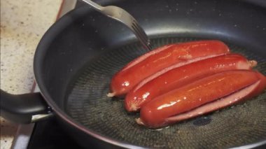 Ev mutfağında sıcak zeytinyağında sosis kızartıyorum. Kızarmış kırmızı sosisleri pişirmek için çatal kullanan bir dişinin kaygan görüntüsü..