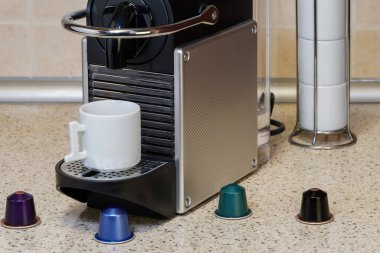 Otomatik bir makinede alüminyum kapsüller kullanarak espresso yaratmak. Kahve makinesinin etrafında metal renkli kapsüller olan mutfak tezgahı ve üzerinde kremalı sıcak kahve yapımında kullanılan beyaz bir fincan..