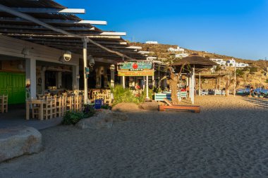 Mykonos, Yunanistan Paradise Plaj Barı 'nda restoran tesisleri var. Cycladic adalarındaki Tropicana kompleksinin akşam manzarası kumun yanındaki boş ahşap sandalye ve masalarla dolu değil.