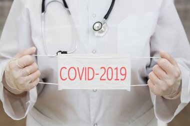 Boynunda fonendoskop, eldivenler ve elinde tıbbi maske olan beyaz önlüklü bir kadın doktor. Maskedeki yazı covid-2019 virüsü hakkında uyarıyor.