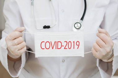 Boynunda fonendoskop, eldivenler ve elinde tıbbi maske olan beyaz önlüklü bir kadın doktor. Maskedeki yazı covid-2019 virüsü hakkında uyarıyor.