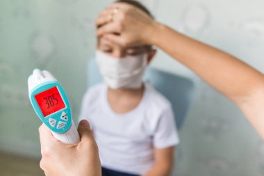 Doktor kız, tıbbi maskeli bir çocuğun vücut ısısını elektronik termometreyle ölçüyor. Çocuktaki virüs yüzünden yüksek ateş..