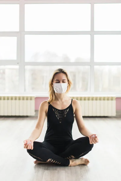 A girl in a medical mask does yoga in a closed Studio in sports clothes. Quiet meditation in the face of a virus epidemic.