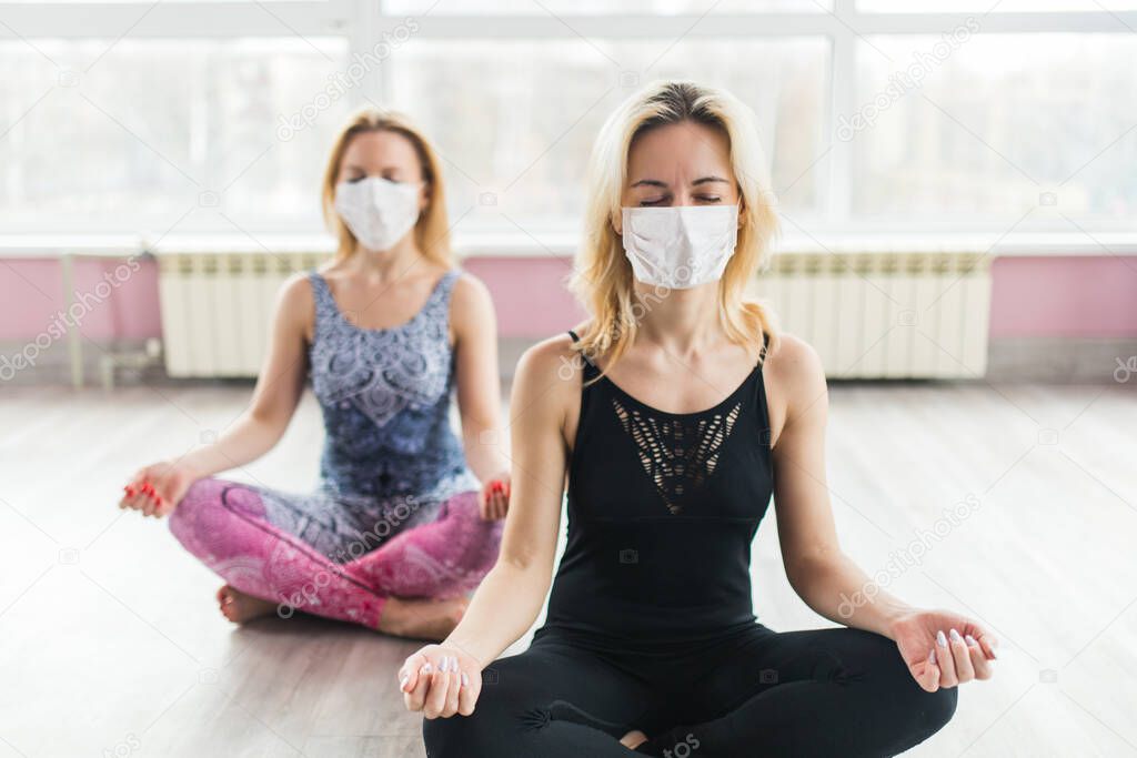 Two girls in medical masks do yoga in a closed Studio in sports clothes. Quiet meditation in the face of a virus epidemic. Group meditation