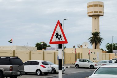 BAE 'de park halindeki araba ve camilerin arka planına karşı bir yol tabelası. Müslüman bir ülkede, yayaların geleneksel kıyafetlerle geçişini gösteren bir tabela. Müslümanlar için yol işareti