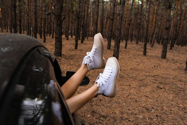 Le gambe di una ragazza in stivali bianchi sporgono da un'auto in una foresta di conifere. Turismo concetto di turismo. Foresta di conifere . — Foto Stock