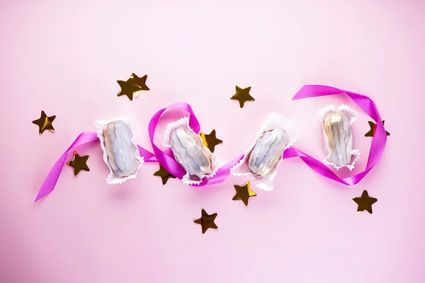 Eclairs with confetti in the form of stars and a purple ribbon on a pink background, flat lay. French dessert. — Stock Photo, Image