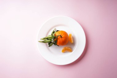 Yeşil dallı bir Mandarin ve pembe arka planda beyaz bir tabakta dilimler. Düz yatıyordu.