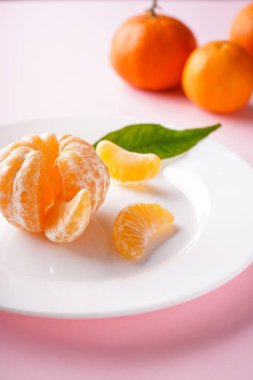 Yeşil dallı mandalinalar ve pembe arka planda beyaz bir tabakta dilimler. Yakın plan..