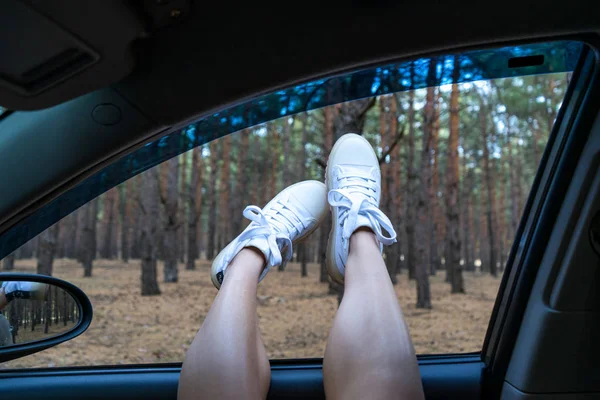 Genuss, Erholung im Wald, fauler Sonntag, Tourismus, Reisen, Freizeit, Erholung, das Konzept des Glücks. Beine eines jungen Mädchens in weißen Stiefeln in einem Autofenster vor dem Hintergrund eines Nadelwaldes. — Stockfoto