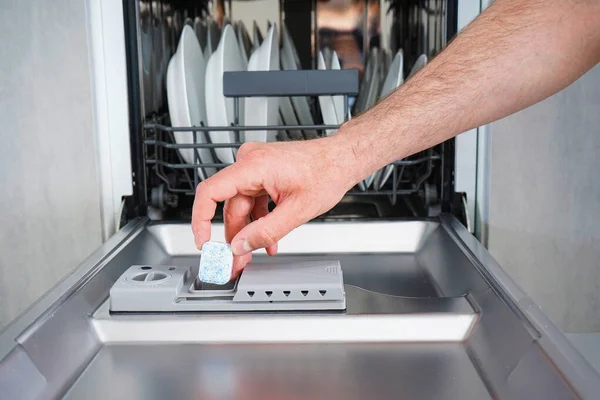 A man hand holding dishwasher detergent tablet. Putting tab into dishwasher, close up.  Dishwasher machine full loaded.