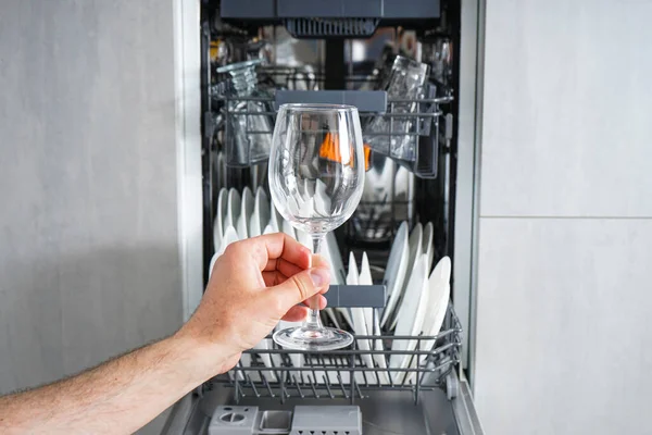 A clean glass in his hand against the background of a loaded dishwasher in the kitchen. Front view.