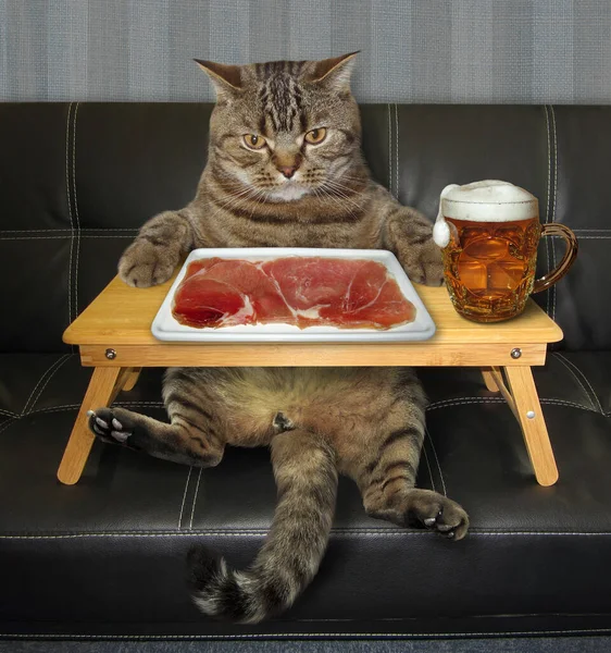 The beige cat is eating a slice of meat which is on a square plate and drinking beer on a black leather sofa. His food is on a wooden bed tray.