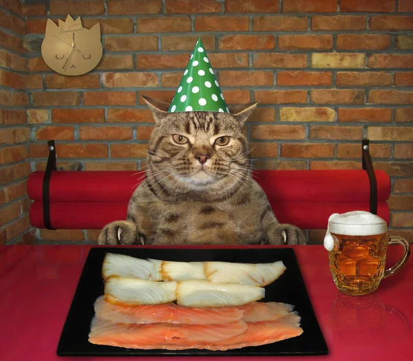 The beige cat in a green birthday hat is eating slices of smoked fish from a black square plate at a table in a restaurant.