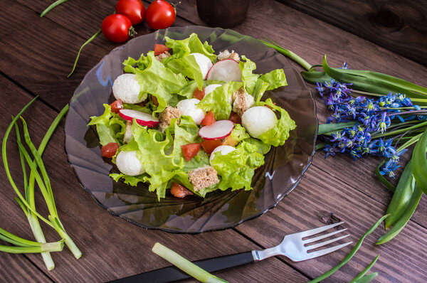 Salad of lettuce, radishes, crackers, tomato and cheese on a transparent dark Circassian plate. Wooden table