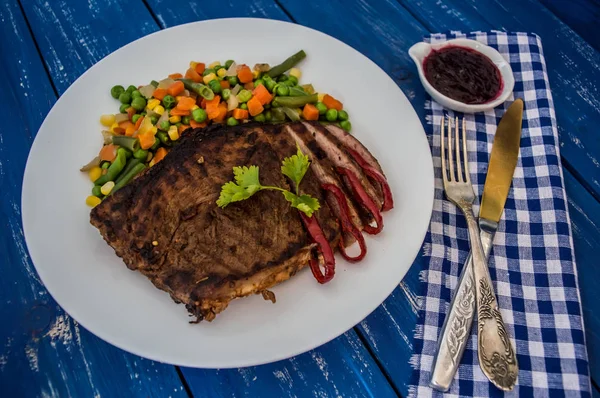 Grillad biff med ångkokta grönsaker på en vit platta. blå tabell. Ovanifrån — Stockfoto