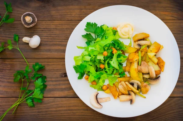 Smażone ziemniaki zapiekane z grzybami na białym talerzu i zielona sałata na tle drewniany stół. Podłoże drewniane. Widok z góry. Szczelnie-do góry — Zdjęcie stockowe